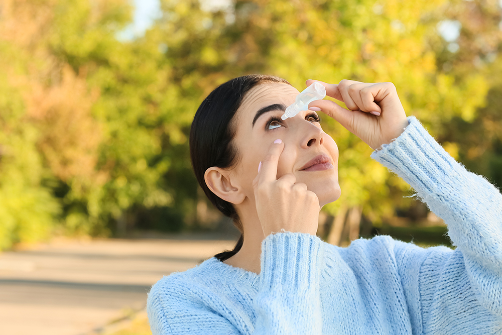 A woman putting eye drops in while standing outside.