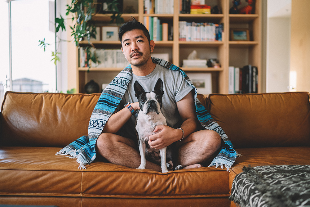 a man sitting on a brown couch with his dog, covered by a blanket