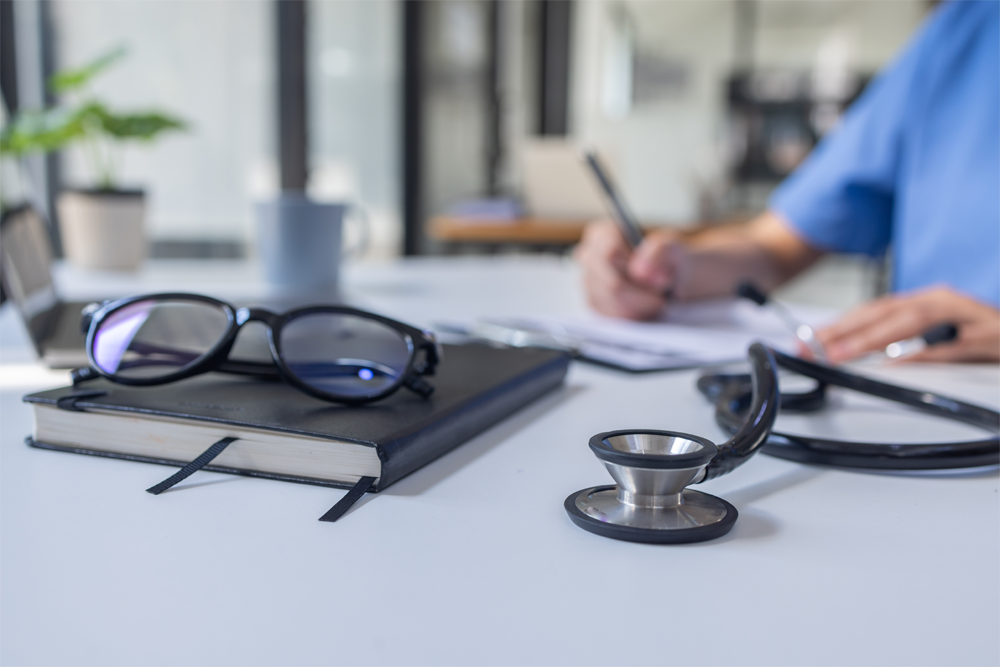 An up-close image of a stethoscope, a notebook, and glasses, with a doctor filling out paperwork in the background.