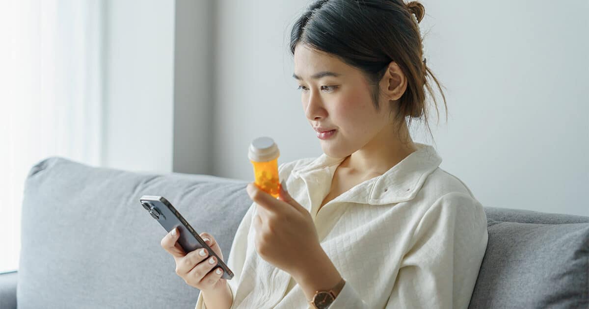 A woman in a white shirt sits on a couch while holding her smartphone in one hand and a pill bottle in the other.