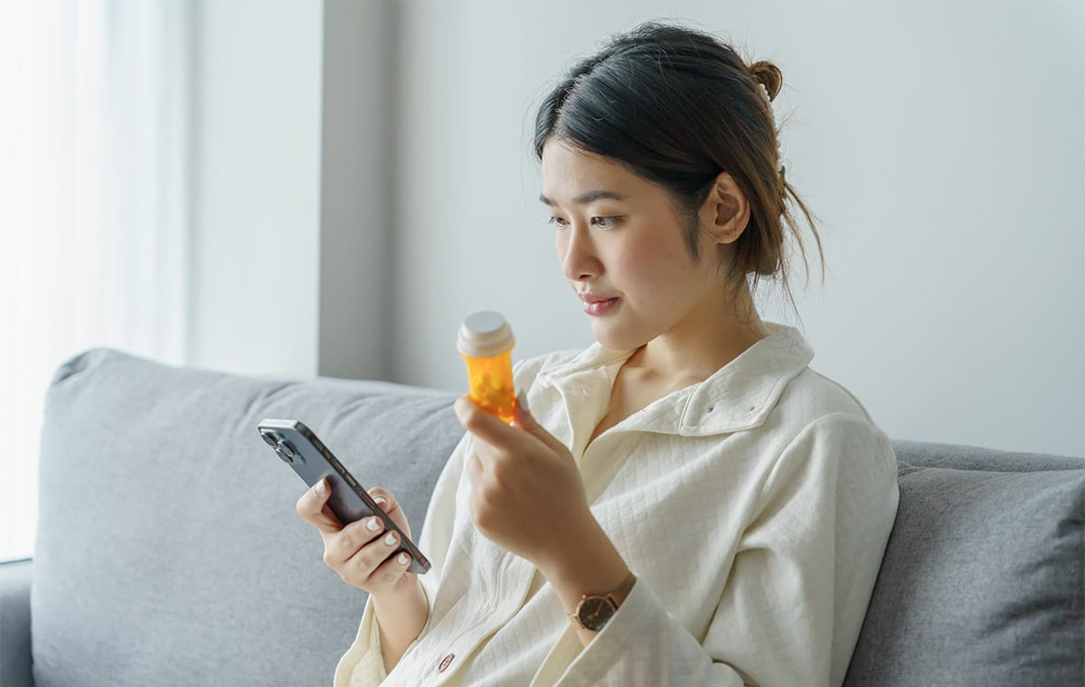 A woman in a white shirt sits on a couch while holding her smartphone in one hand and a pill bottle in the other.