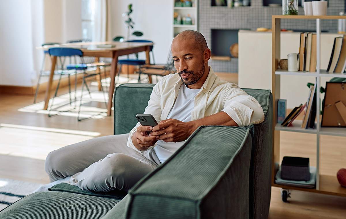 An individual comparing TelyRx vs. Hims on their phone while sitting on a green corduroy couch in a modern, open living space.