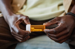 A man sits on his bed and holds a small bottle of prescription medication. The bottle has several oblong pills inside.