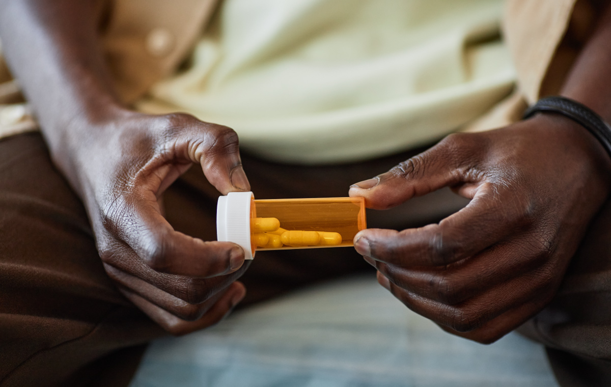 A man sits on his bed and holds a small bottle of prescription medication. The bottle has several oblong pills inside.